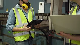 The image displays an industrial worker checking data on a tablet in a factory setting. 