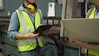 The image displays an industrial worker checking data on a tablet in a factory setting. 