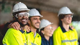 a group of diverse workers in high-visibility vests and safety helmets smiling and embracing in what appears to be a workplace setting