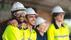 a group of diverse workers in high-visibility vests and safety helmets smiling and embracing in what appears to be a workplace setting