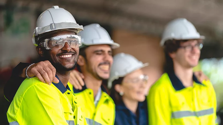 a group of diverse workers in high-visibility vests and safety helmets smiling and embracing in what appears to be a workplace setting